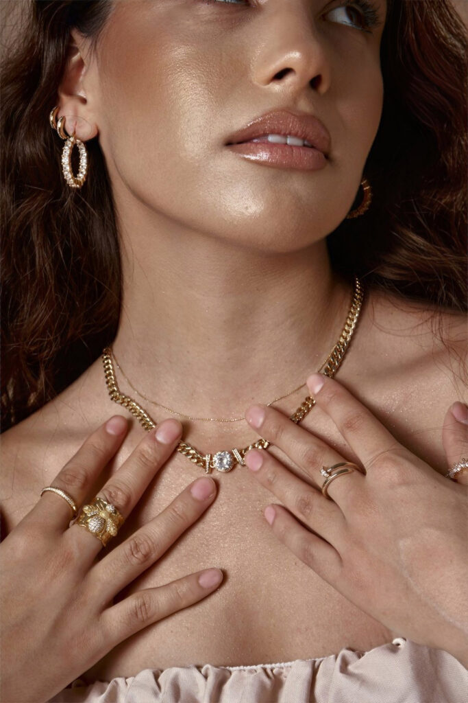 Abril Escalante showcases her elegant jewelry including gold hoop earrings a thick gold chain necklace and multiple rings against a soft beige background.